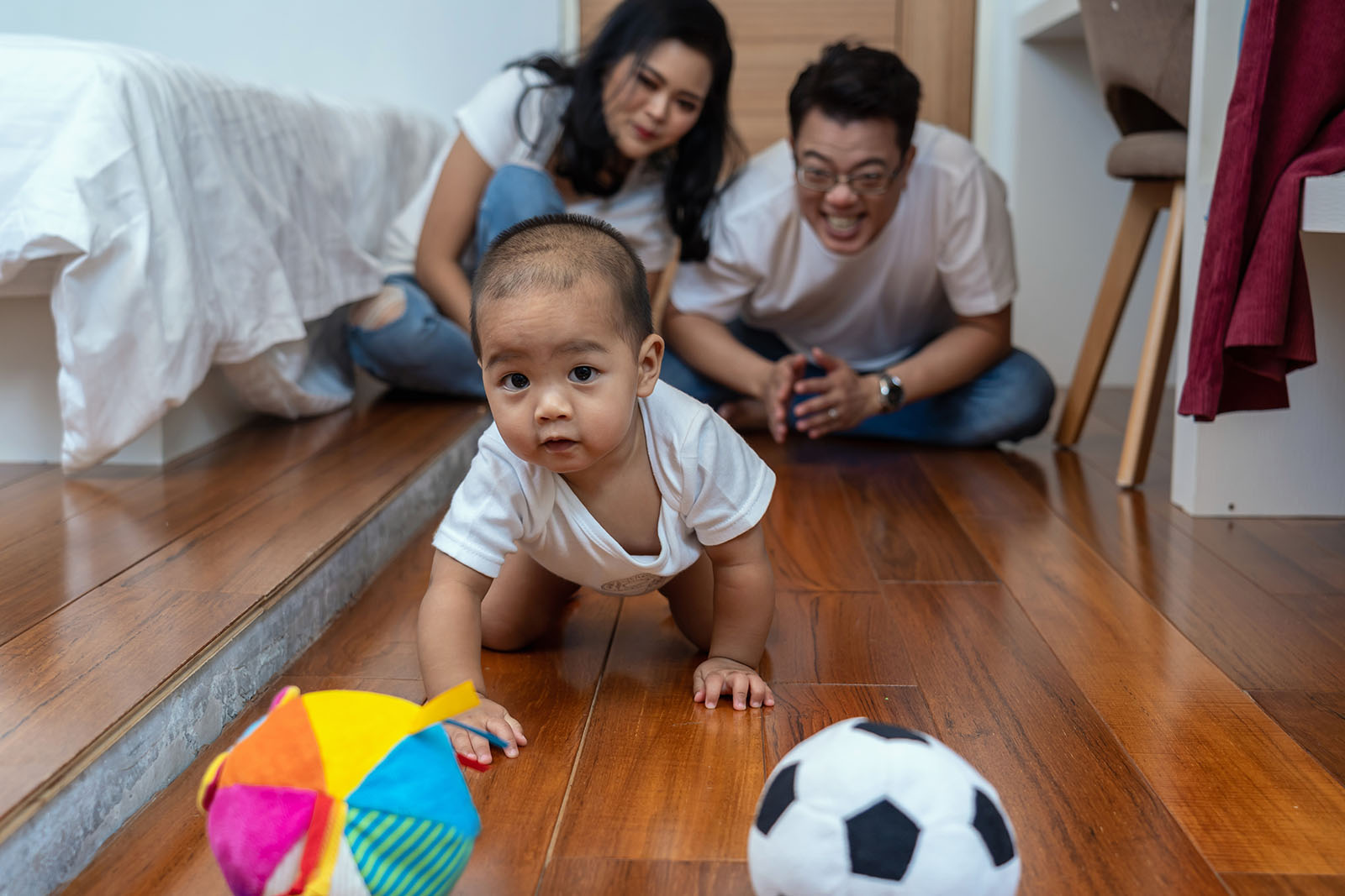 Baby boy crawls and plays with balls on the wooden floor, with father and mother smiling in the background.